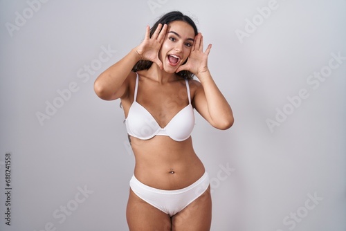 Young hispanic woman wearing white lingerie smiling cheerful playing peek a boo with hands showing face. surprised and exited