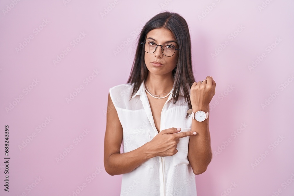 Brunette young woman standing over pink background wearing glasses in hurry pointing to watch time, impatience, looking at the camera with relaxed expression Brunette young woman standing over pink background wearing glasses in hurry pointing to watch time, impatience, looking at the camera with relaxed expression