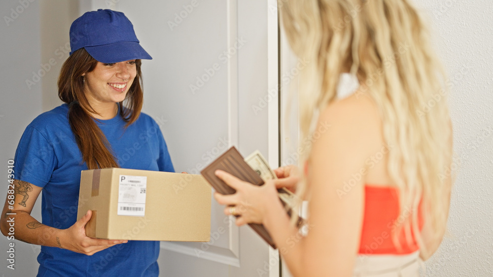 Young beautiful hispanic woman delivery woman giving package to client paying dollars at home Young beautiful hispanic woman delivery woman giving package to client paying dollars at home
