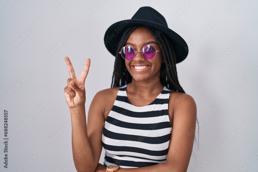 Young african american with braids wearing hat and sunglasses smiling with happy face winking at the camera doing victory sign with fingers. number two. Young african american with braids wearing hat and sunglasses smiling with happy face winking at the camera doing victory sign with fingers. number two.