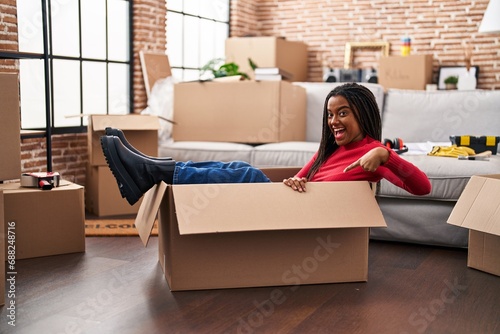 Young african american with braids moving to a new home inside of a cardboard box smiling happy pointing with hand and finger © Krakenimages.com