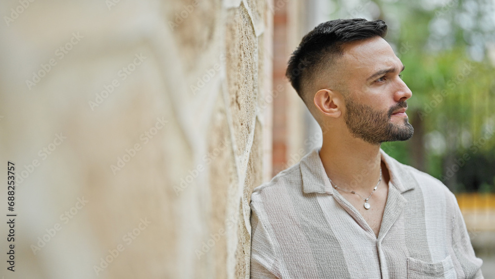 Young hispanic man looking to the side with serious expression leaning on wall at street Young hispanic man looking to the side with serious expression leaning on wall at street