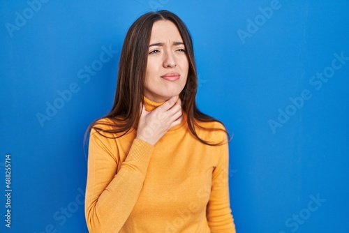Young brunette woman standing over blue background touching painful neck, sore throat for flu, clod and infection