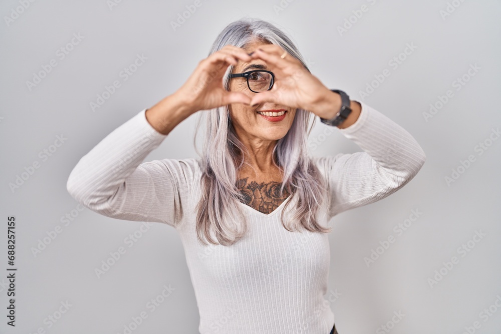Middle age woman with grey hair standing over white background doing heart shape with hand and fingers smiling looking through sign Middle age woman with grey hair standing over white background doing heart shape with hand and fingers smiling looking through sign