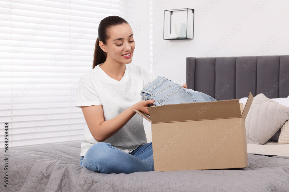 Happy young woman taking jeans out of box on bed at home. Online shopping Happy young woman taking jeans out of box on bed at home. Online shopping