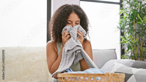 Beautiful young hispanic woman smiling joyfully, smelling freshly-washed clean clothes on sofa at home, indoors, indulging in the fragrant aroma of domestic chores photo