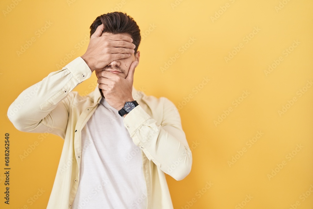 Young hispanic man standing over yellow background covering eyes and mouth with hands, surprised and shocked. hiding emotion Young hispanic man standing over yellow background covering eyes and mouth with hands, surprised and shocked. hiding emotion