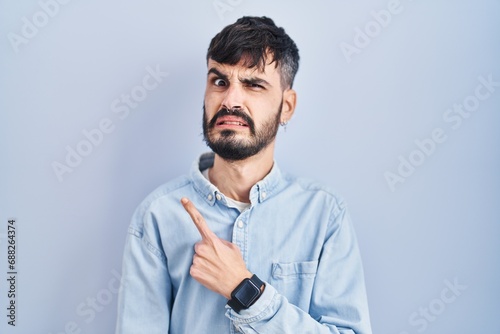 Young hispanic man with beard standing over blue background pointing aside worried and nervous with forefinger, concerned and surprised expression