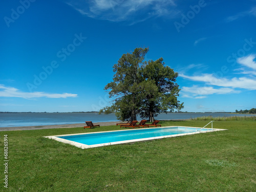 Lawn terrace with swimming pool and deckchairs under a tree for relaxation at the shore of a lake