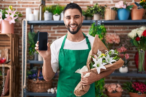 Hispanic young man working at florist shop showing smartphone screen smiling and laughing hard out loud because funny crazy joke.