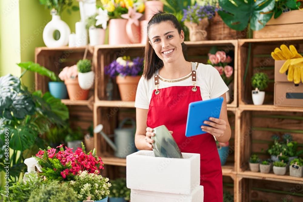 Young beautiful hispanic woman florist unpacking plant jar using touchpad at flower shop