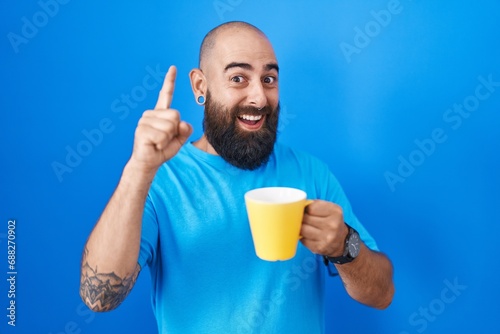Young hispanic man with beard and tattoos drinking a cup of coffee pointing finger up with successful idea. exited and happy. number one. © Krakenimages.com