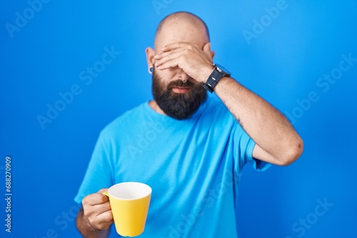 Young hispanic man with beard and tattoos drinking a cup of coffee covering eyes with hand, looking serious and sad. sightless, hiding and rejection concept © Krakenimages.com