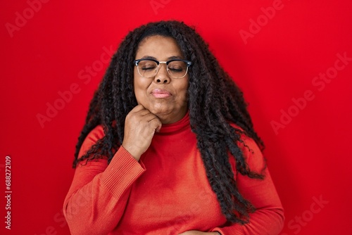 Plus size hispanic woman standing over red background with hand on chin thinking about question, pensive expression. smiling with thoughtful face. doubt concept. © Krakenimages.com