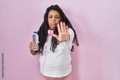 Plus size hispanic woman wearing sportswear and headphones doing stop sing with palm of the hand. warning expression with negative and serious gesture on the face.