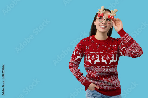 Happy young woman in Christmas sweater and funny glasses on light blue background. Space for text