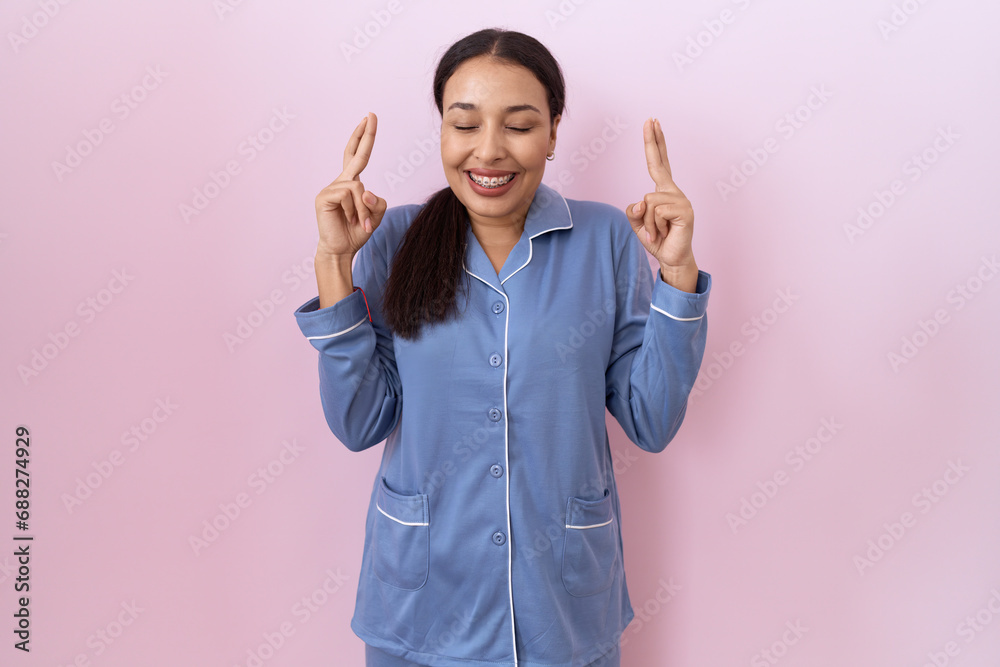 Young arab woman wearing blue pajama gesturing finger crossed smiling with hope and eyes closed. luck and superstitious concept.