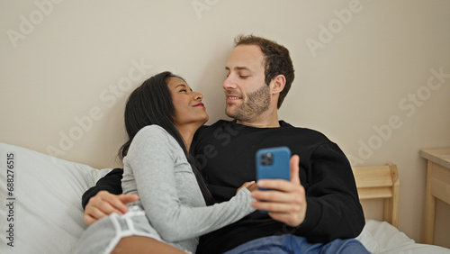 Beautiful couple lying on bed using smartphone smiling at bedroom