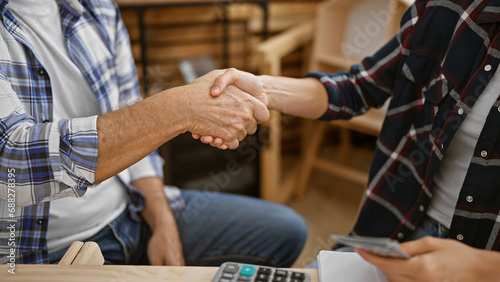 Two hearty carpenters, handing over dollars, seal a woodwork agreement with a firm handshake at their cozy indoor carpentry workplace © Krakenimages.com