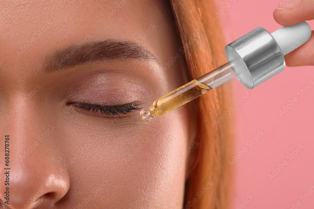 Obraz premium Beautiful young woman applying cosmetic serum onto her face on pink background, closeup