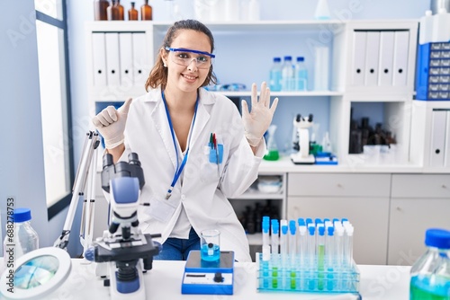 Young hispanic woman working at scientist laboratory showing and pointing up with fingers number six while smiling confident and happy.