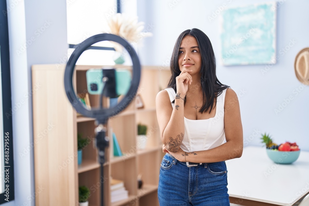 Brunette woman recording vlog tutorial with smartphone at home thinking concentrated about doubt with finger on chin and looking up wondering Brunette woman recording vlog tutorial with smartphone at home thinking concentrated about doubt with finger on chin and looking up wondering
