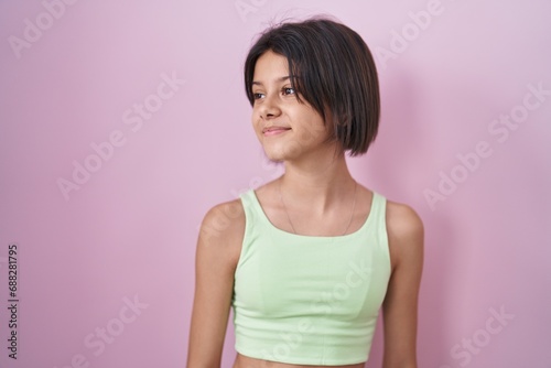 Young girl standing over pink background looking away to side with smile on face  natural expression. laughing confident.