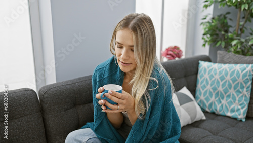 Beautiful young blonde woman wrapped in a cozy blanket at home, warming up with a hot mug of coffee on a cold winter's day