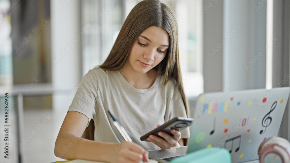 Young beautiful girl student using smartphone taking notes at library