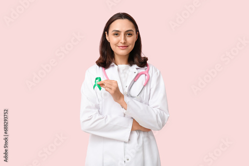 Young female doctor with green ribbon on pink background. Liver cancer awareness concept