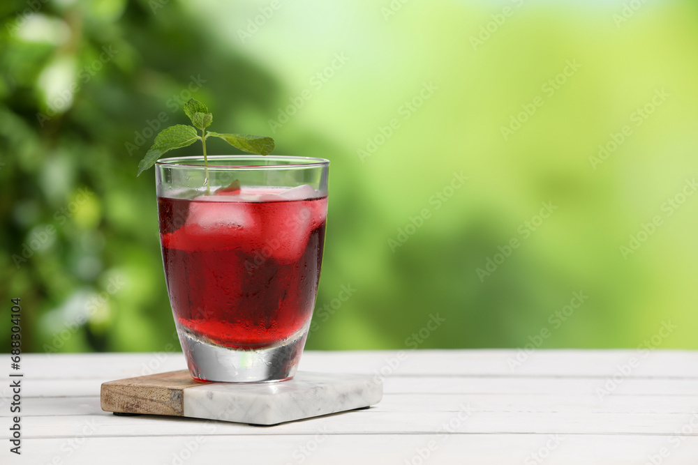Refreshing hibiscus tea with ice cubes and mint in glass on white wooden table against blurred green background. Space for text Refreshing hibiscus tea with ice cubes and mint in glass on white wooden table against blurred green background. Space for text