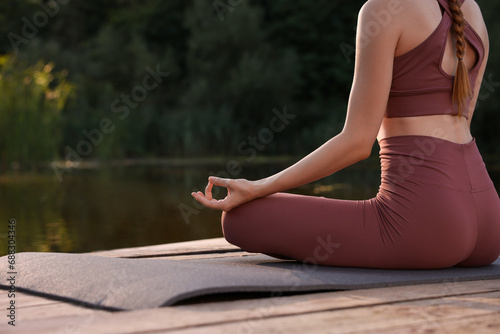 Woman practicing Padmasana on yoga mat on wooden pier near pond, closeup with space for text. Lotus pose