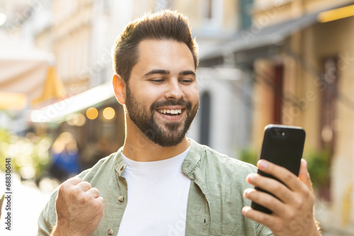 Caucasian young man use mobile smartphone celebrating win good message news, lottery jackpot victory, giveaway online outdoors. Happy adult guy tourist walking in urban city street. Town lifestyles
