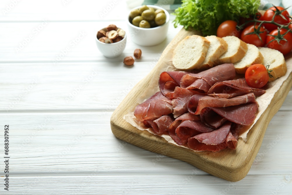 Board with delicious bresaola served with bread and tomato on white wooden table. Space for text