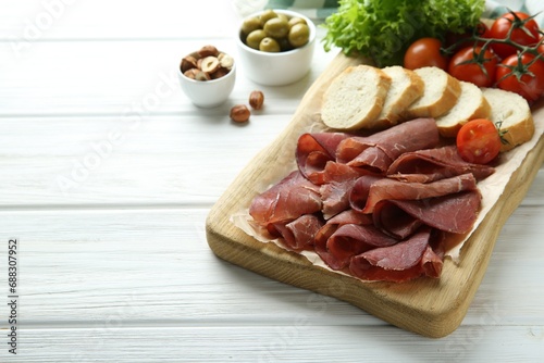 Board with delicious bresaola served with bread and tomato on white wooden table. Space for text