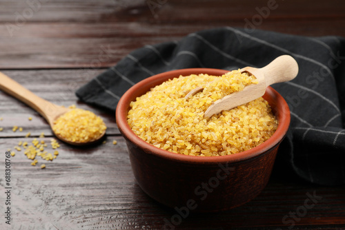 Bowl and spoon with raw bulgur on wooden table, closeup