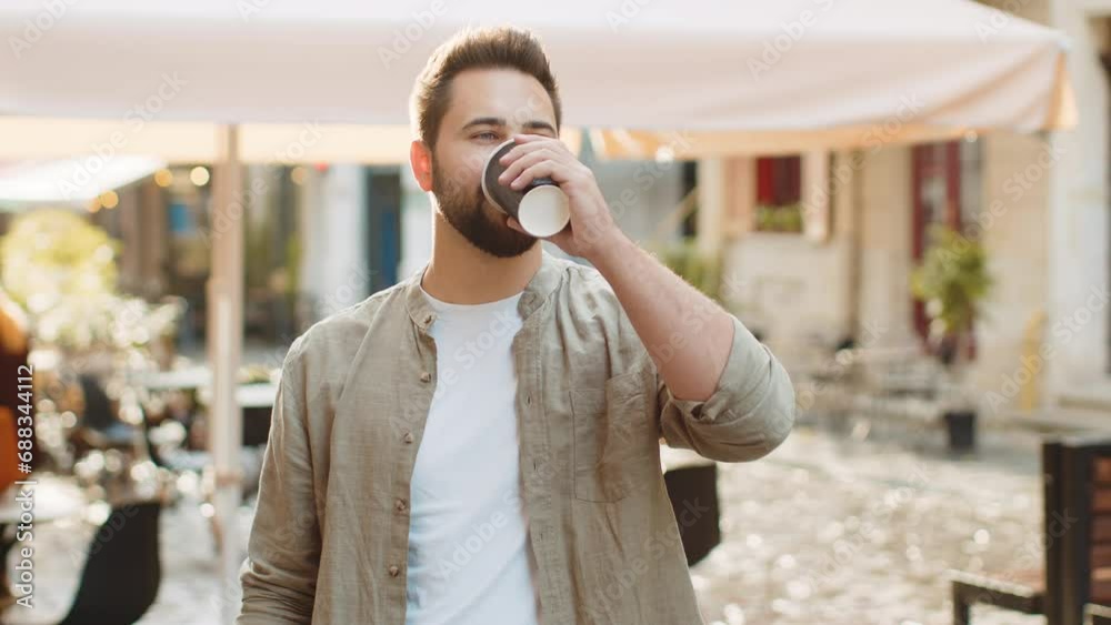 Happy bearded young man enjoying morning coffee hot drink and smiling outdoors. Relaxing, taking a break. Guy walking in urban city sunshine street, drinking coffee to go. Town lifestyles outside