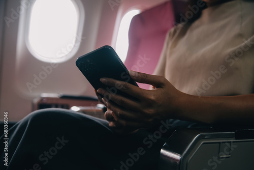 Asian woman passenger sitting in airplane near window and reading news from social networks or using travel applications in smartphone
