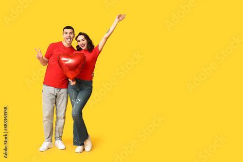 Loving young couple with heart-shaped balloon showing victory gesture on yellow background. Celebration of Saint Valentine's Day
