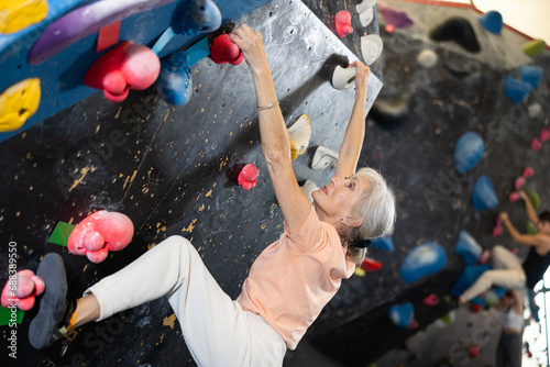 Sporty elderly female exercising in boulder climbing hall without rope, reaching new results, enjoying new challenges in amusement park