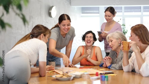 Group of cheerful carefree female colleagues of different ages and nationalities enjoying playing board game around table during work break in office photo