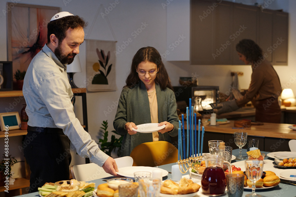 Mature man and his daughter putting plates and other cutlery on dinner table with homemade traditional Jewish food prepared for Hanukkah