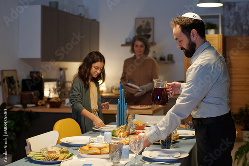 Side view of bearded man in white shirt helping serve dinner table with plates and glass jug of homemade juice for Hanukkah celebration