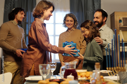 Happy young brunette woman passing packed giftbox to youthful girl over served table while family members giving each other Hanukkah presents photo