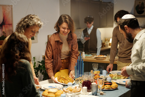 Happy Jewish family putting plates with homemade food on table while serving it for invited guests before Hanukkah dinner photo
