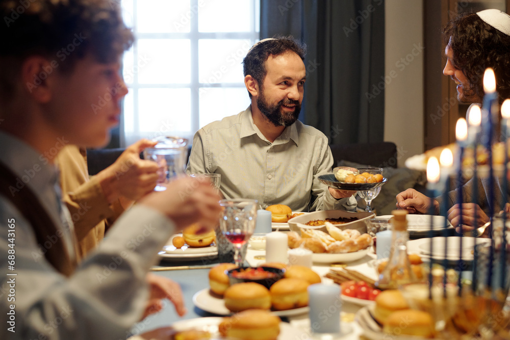 Focus on mature bearded Jewish man offering his son or guest try traditional homemade pastry while holding plate with tasty donuts