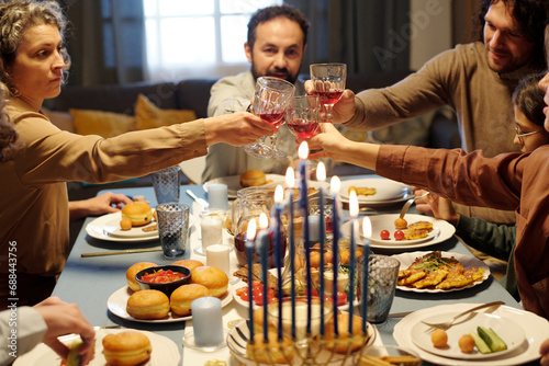 Members of large Jewish family clinking with wineglasses over served table with homemade food while toasting for Hanukkah by dinner photo