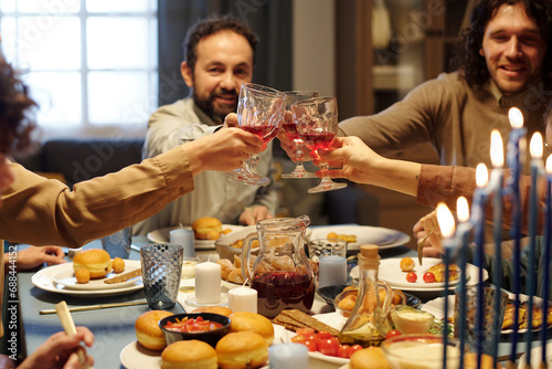 Hands of Jewish family members clinking with glasses of red wine over festive table served with homemade snacks during celebration photo