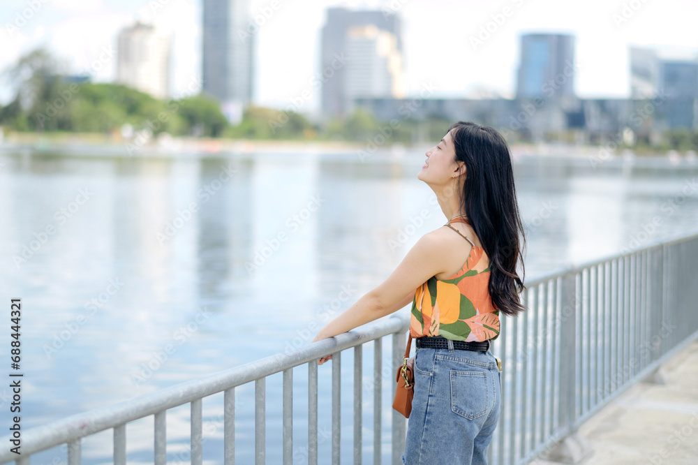 In a vibrant blue jeans and orange shirt, a cheerful lady stands by the lake, creating perfect outdoor portrait.