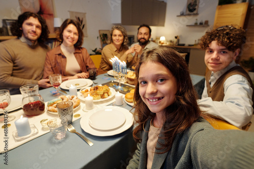 Cute smiling girl taking selfie with members of her family by dinner while sitting by served table with homemade food and drinks